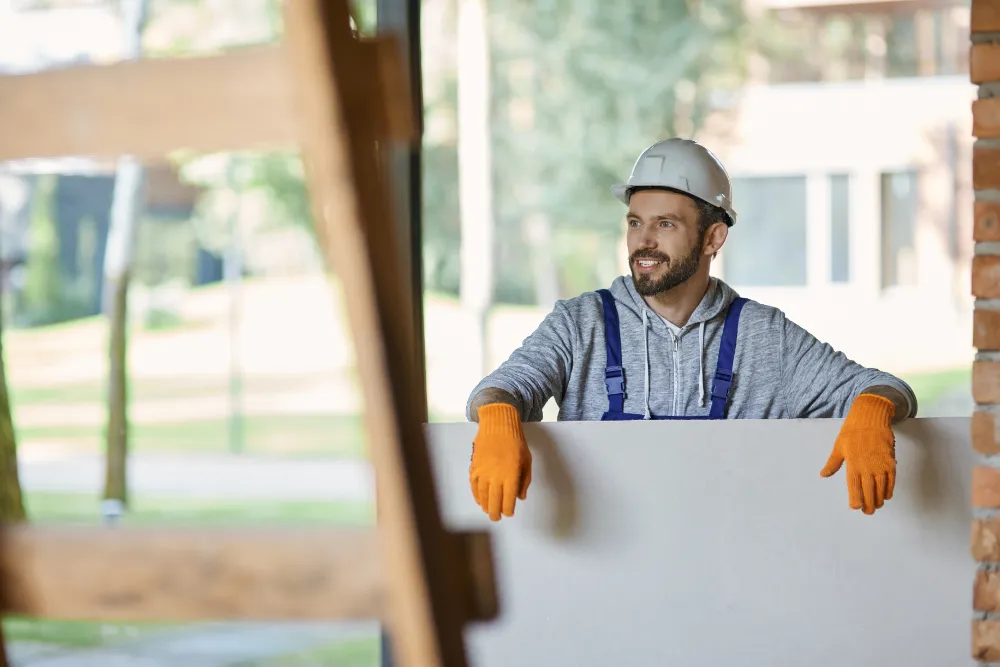 Happy young male builder in hard hat holding drywall while working at construction site